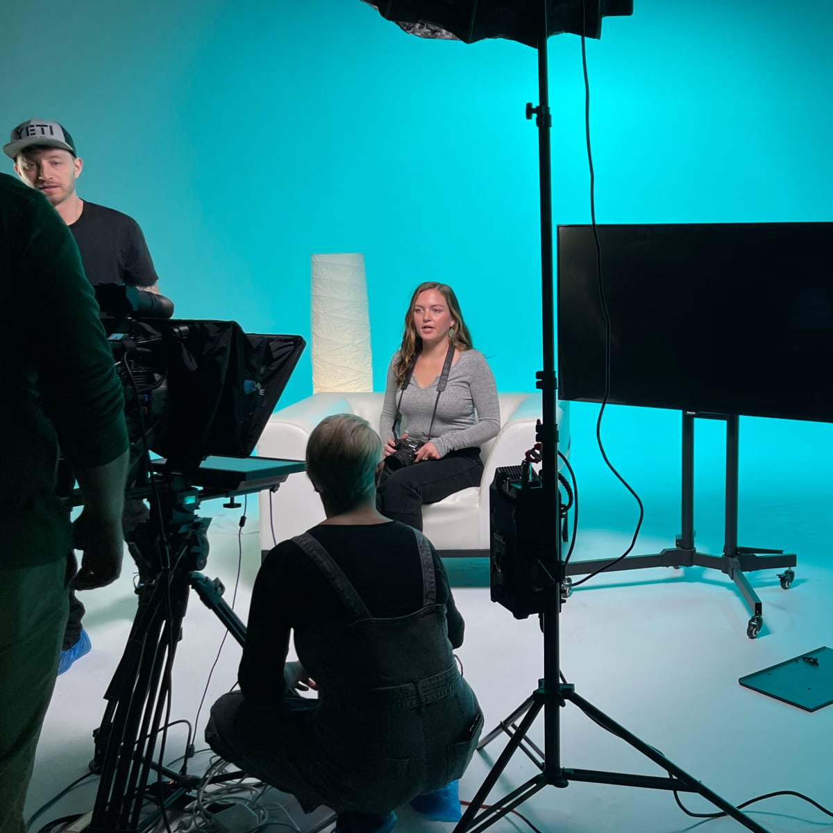 A woman sits on a sofa in front of a large screen, while two crew members prepare equipment on a brightly lit set with a blue backdrop.