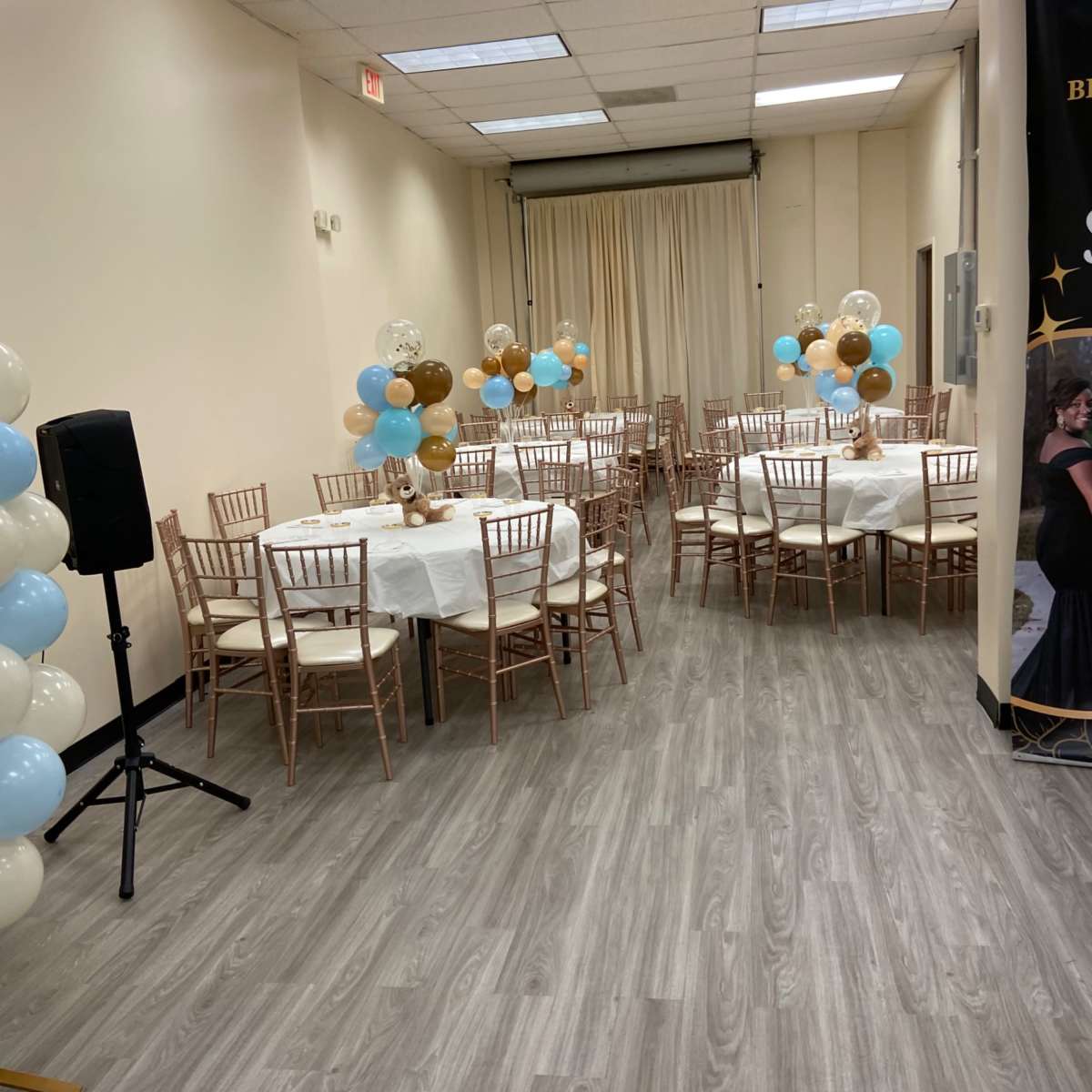 A decorated event space with tables set for a gathering, featuring balloons in blue and gold accents.