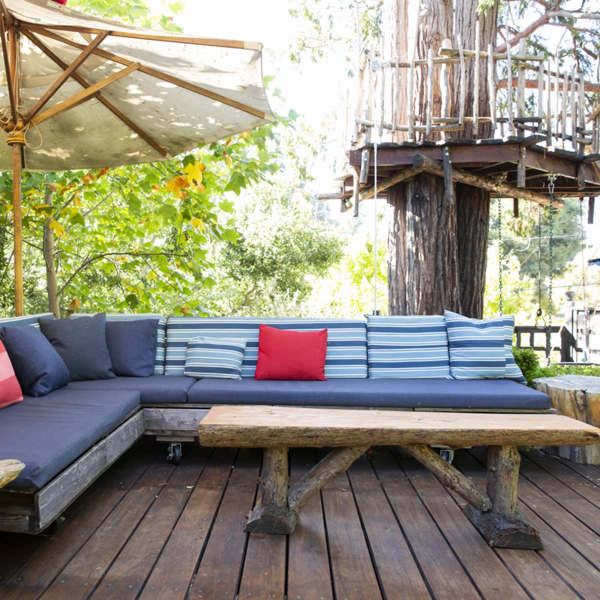 A wooden deck features a sectional sofa with striped cushions, a wooden table, and a large umbrella shaded by trees.