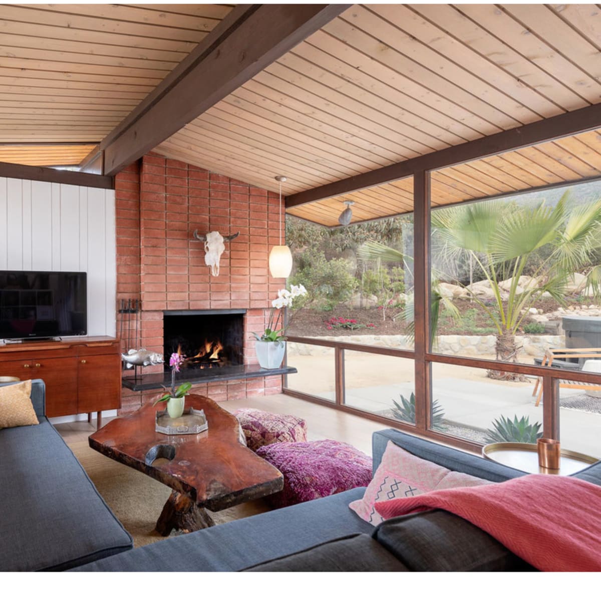 A modern living room features a wood-paneled ceiling, a large sectional sofa, a coffee table made from natural wood, and a fireplace surrounded by brick.