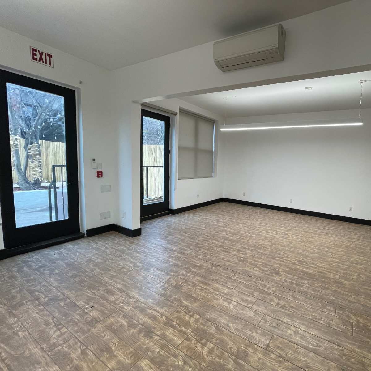 An empty room with wood-like flooring, a large window, and an exit door leading outside.