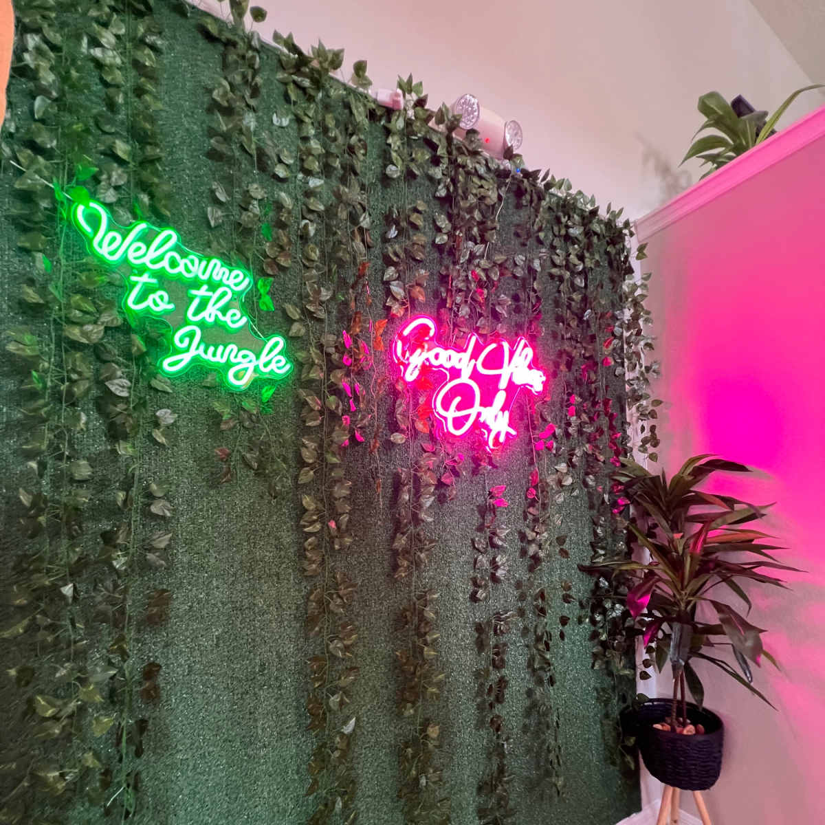 A wall covered in greenery with neon signs that read "Welcome to the Jungle" and "Good Vibes Only," alongside a plant in a basket on a stand.