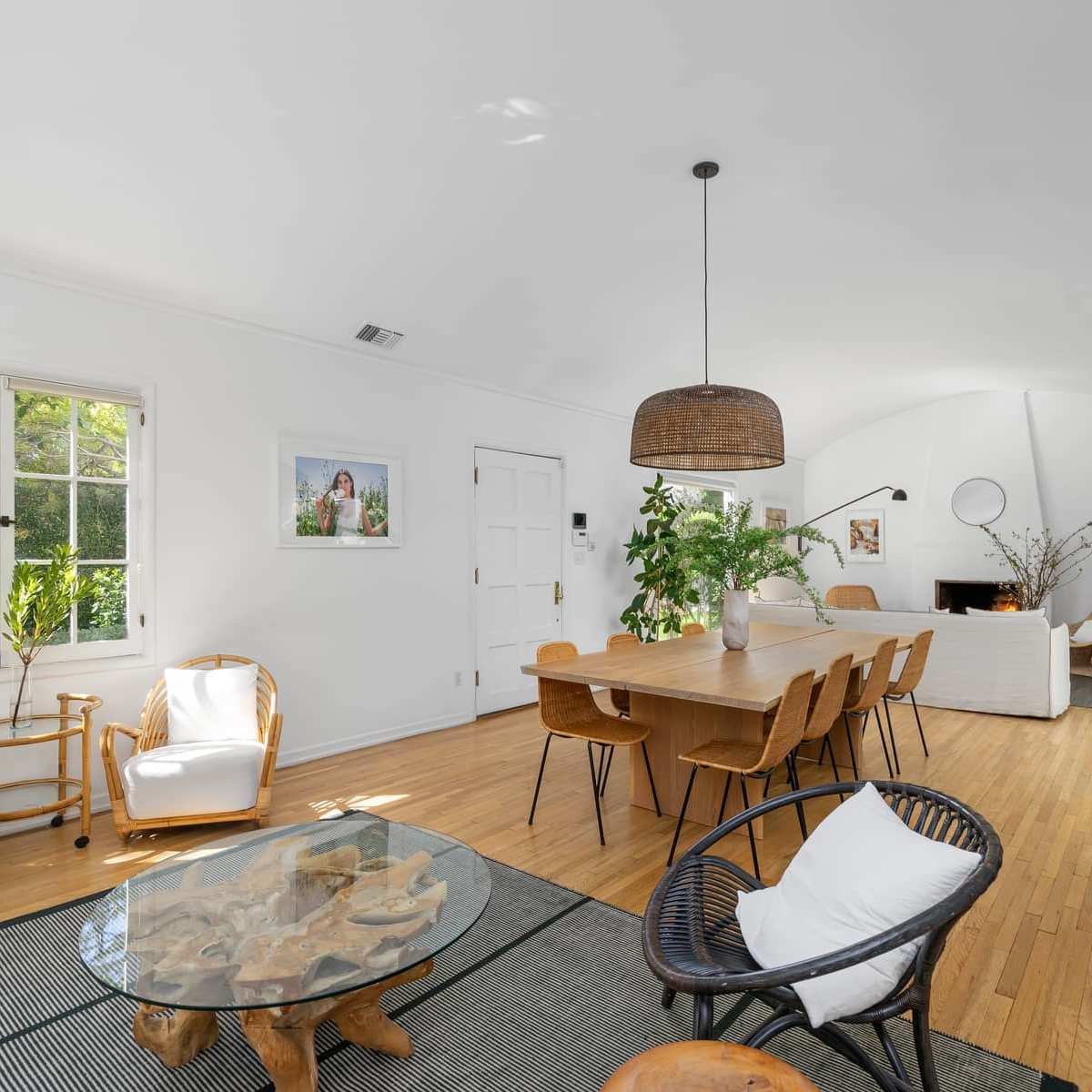 A spacious, sunlit living area features a wooden dining table surrounded by chairs, a glass coffee table, and various indoor plants.