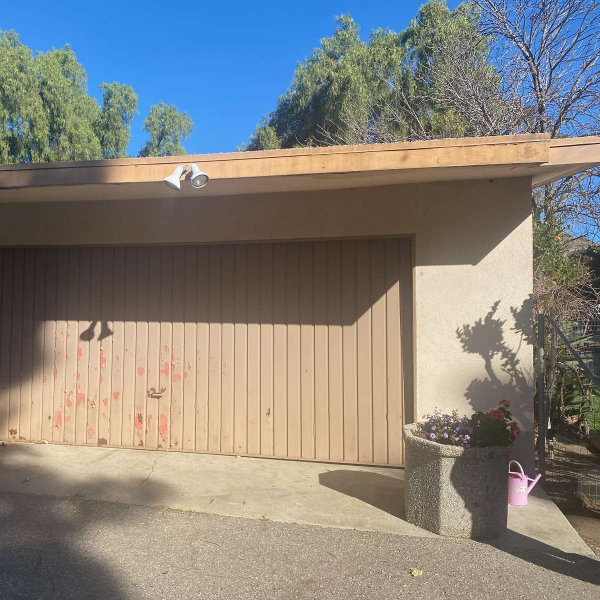 A garage with a closed door, featuring weathered paint and a flower planter nearby, is set against a clear blue sky.