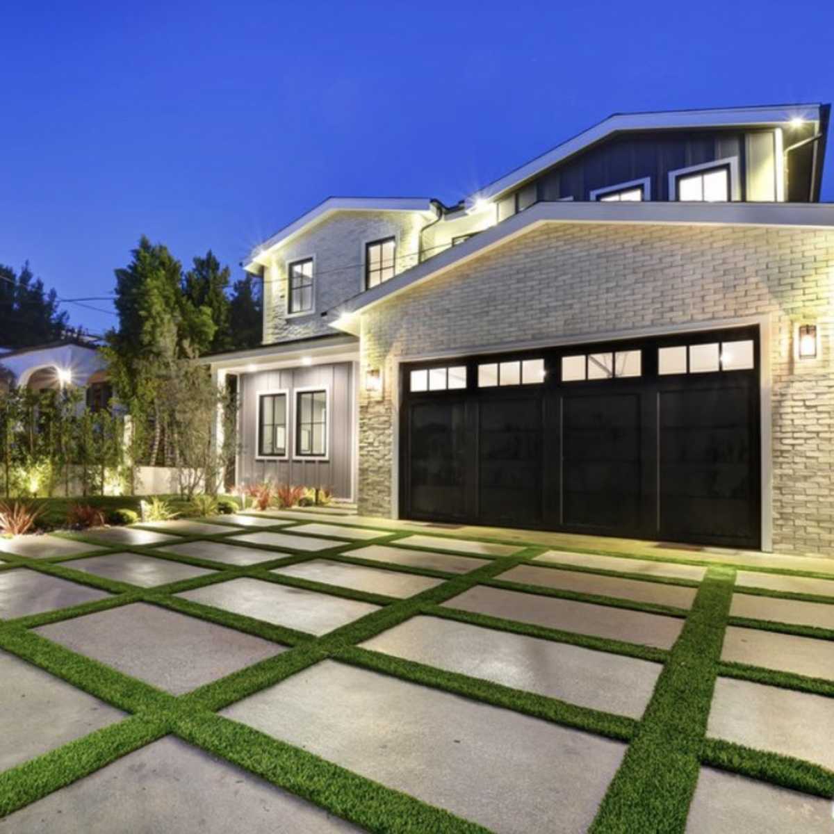 A modern house with a well-lit facade, featuring a manicured lawn and a geometric concrete driveway with grass strips.