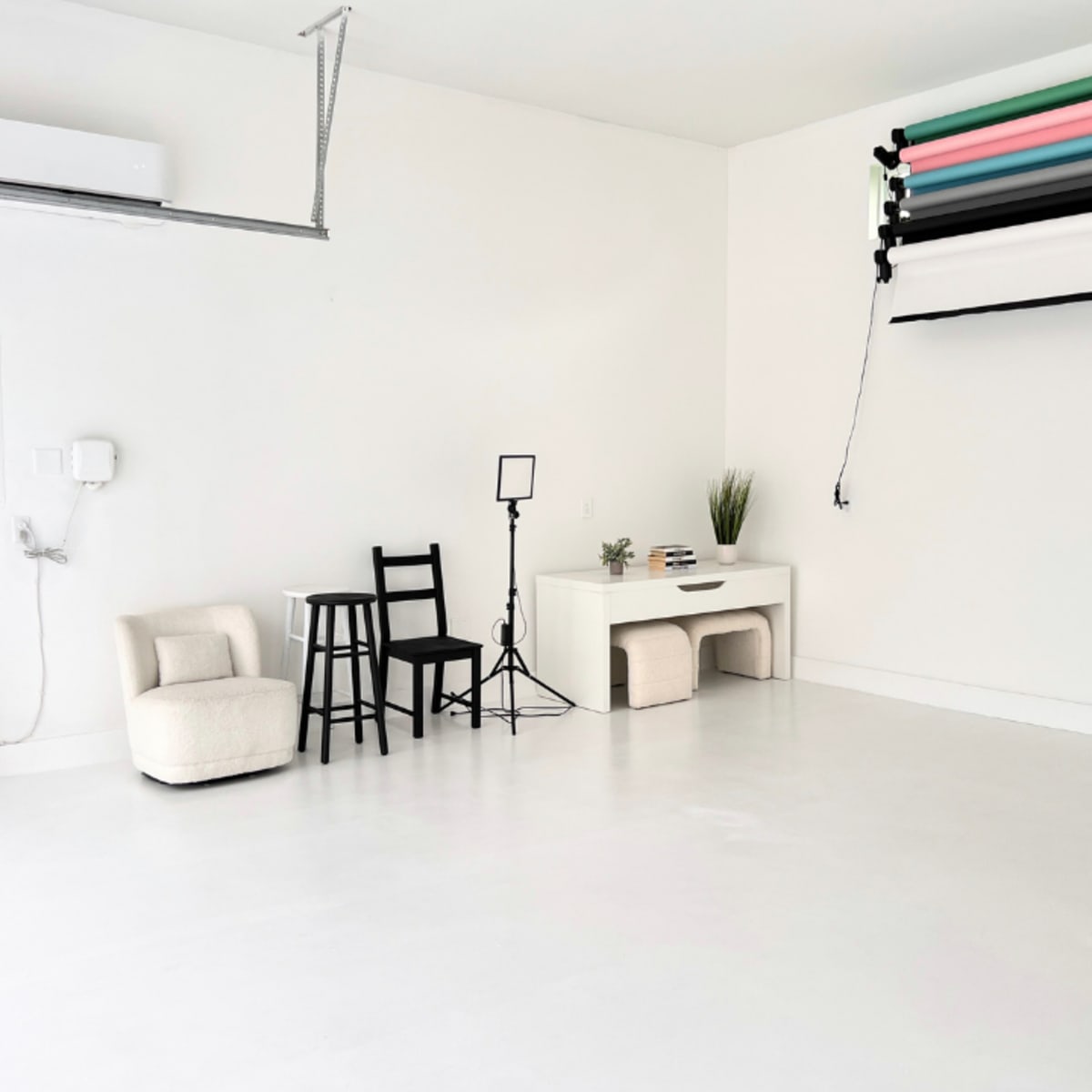 A minimalist studio space with white walls and a smooth floor, featuring a chair, a small table with a plant, a light stand, and a roll of backdrops mounted on the wall.