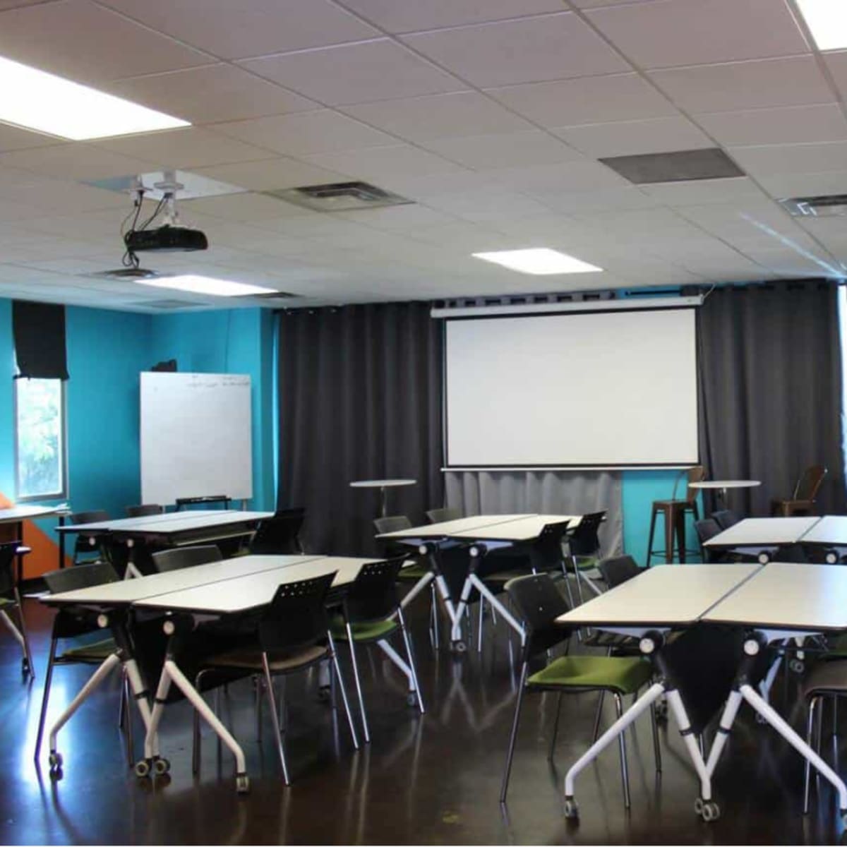 A modern classroom setup with several tables and chairs arranged facing a projector screen, surrounded by colorful walls and curtains.