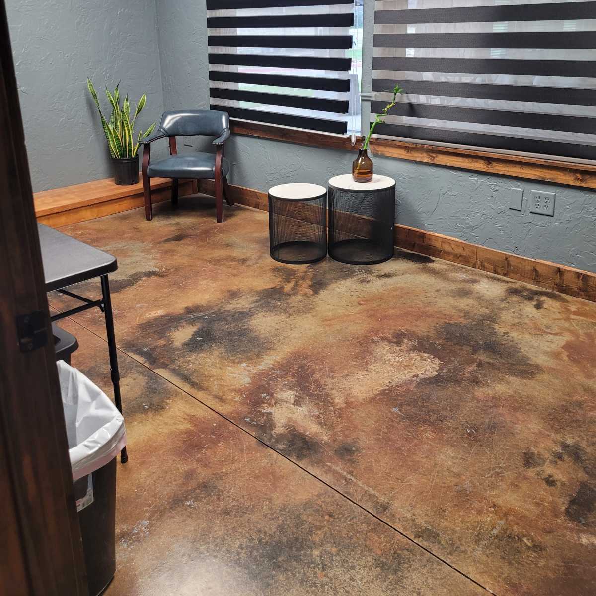 A small room with concrete floors, two striped window shades, a chair, a small table, and a plant in the corner.
