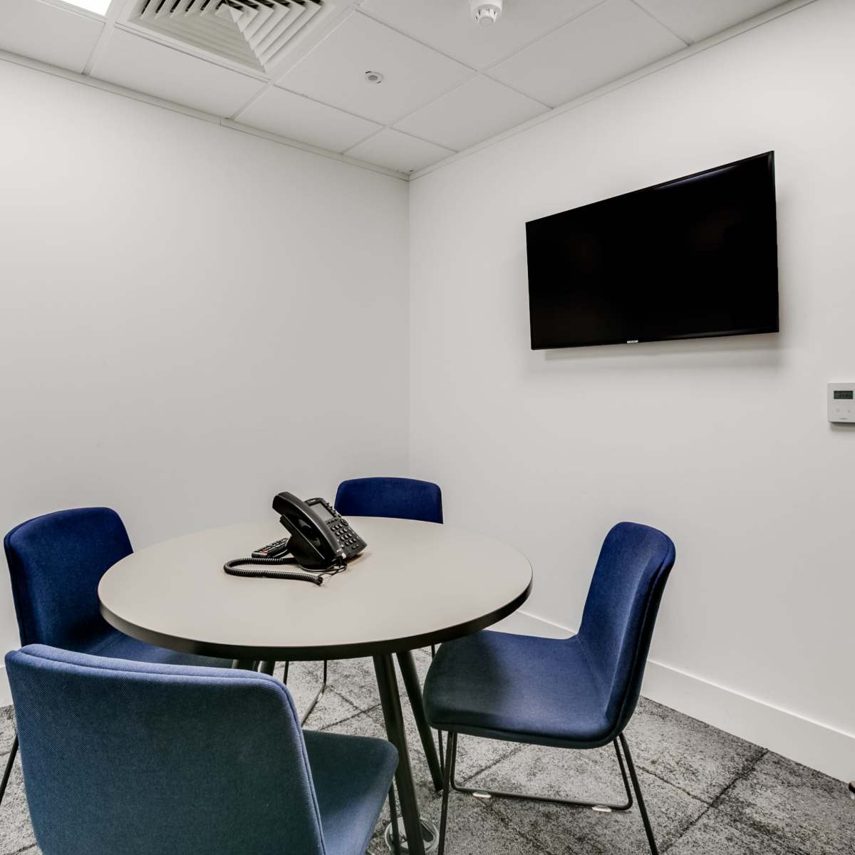 A small meeting room features a round table with four blue chairs and a telephone, along with a mounted television on the wall.