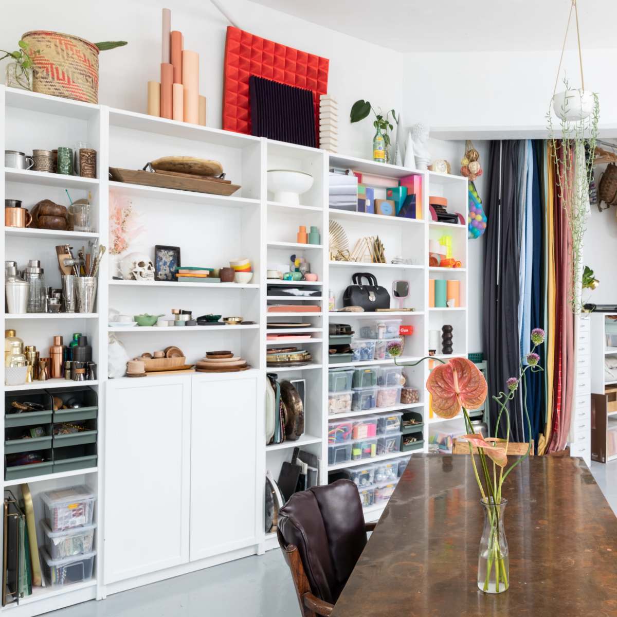 A bright room featuring a wooden dining table surrounded by chairs, with shelves filled with various knickknacks and organized storage units on one side.