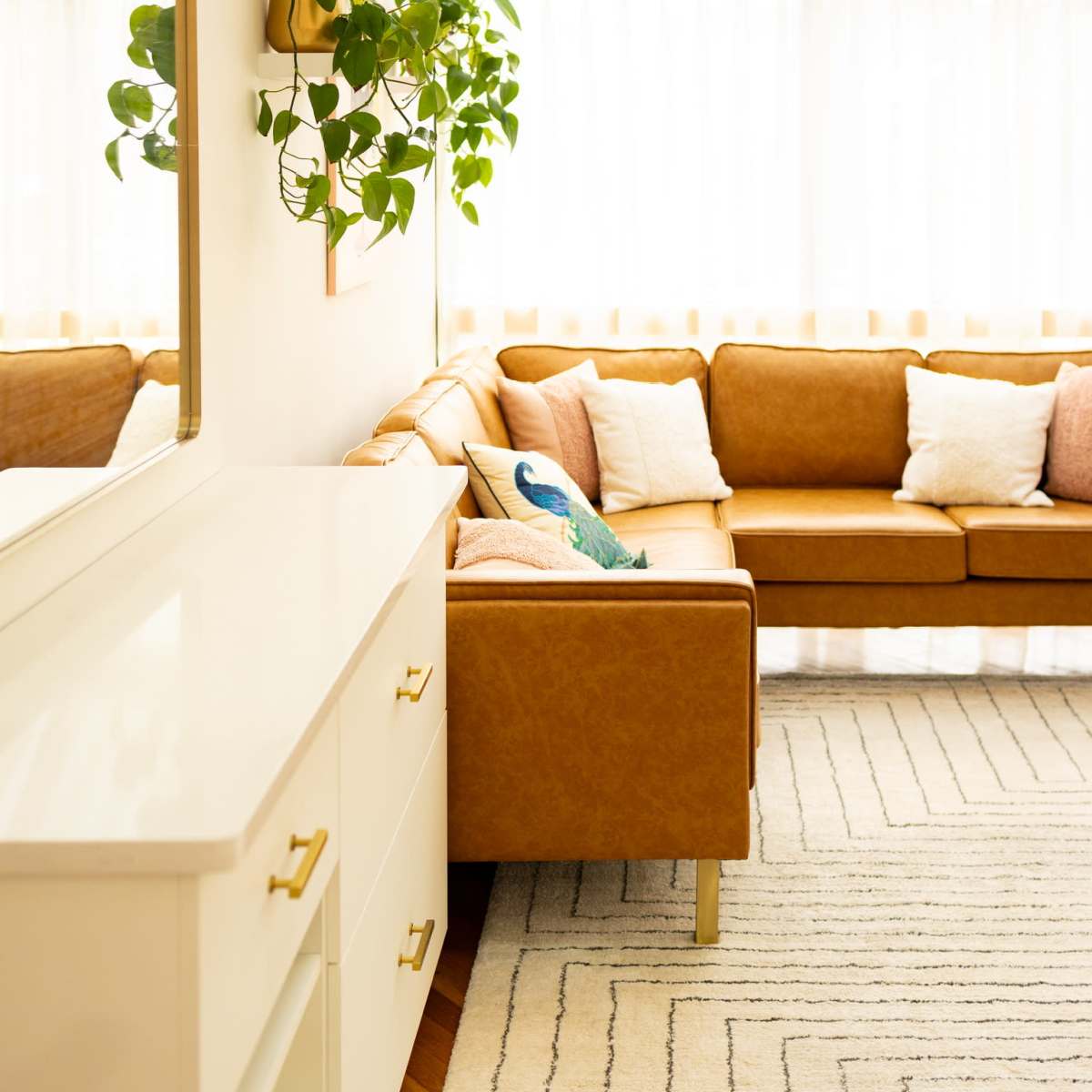 A well-lit living room features a brown leather sofa, a white dresser, and a plant hanging on the wall.