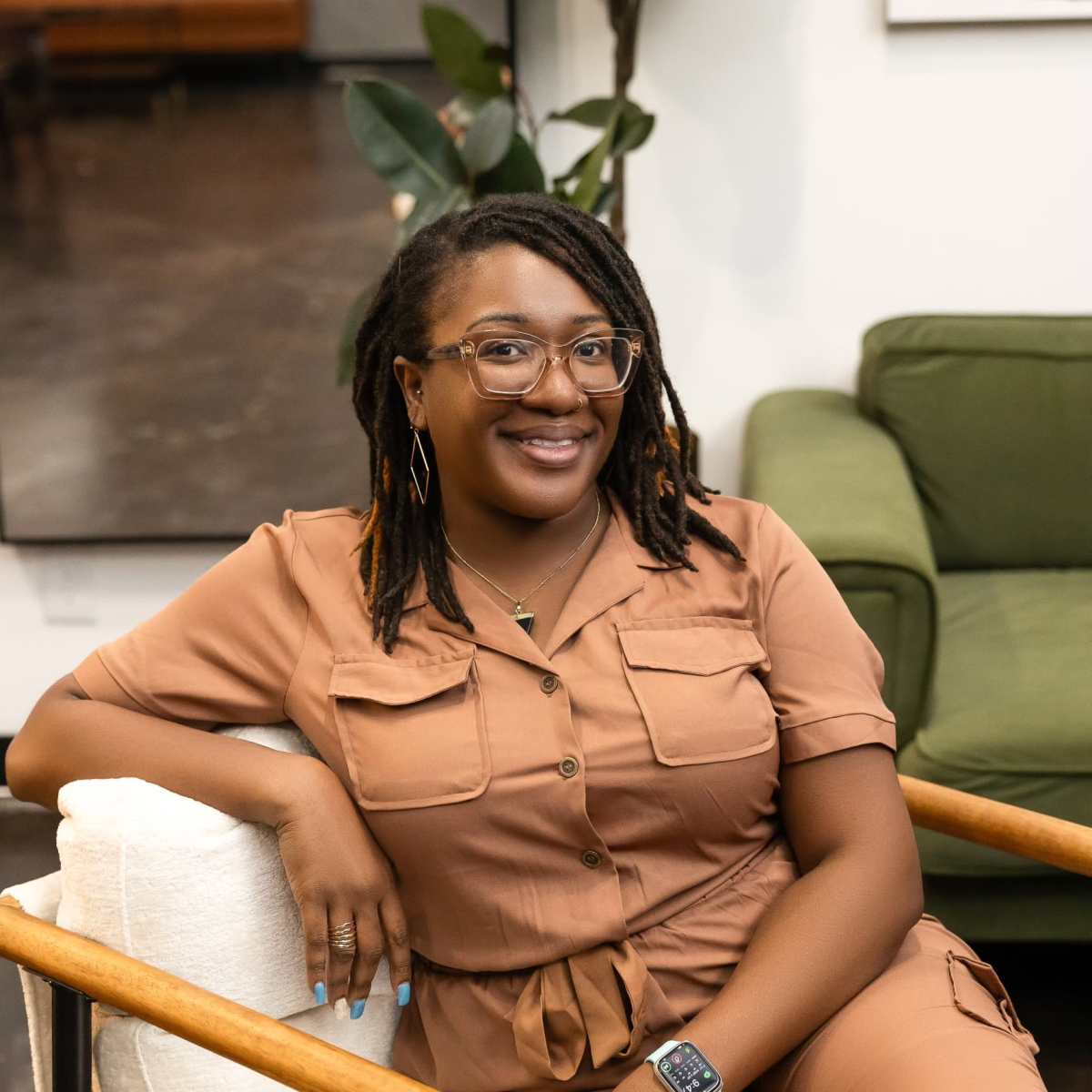 A person with glasses and dreadlocks sits comfortably in a modern chair, smiling at the camera in a stylish indoor space.