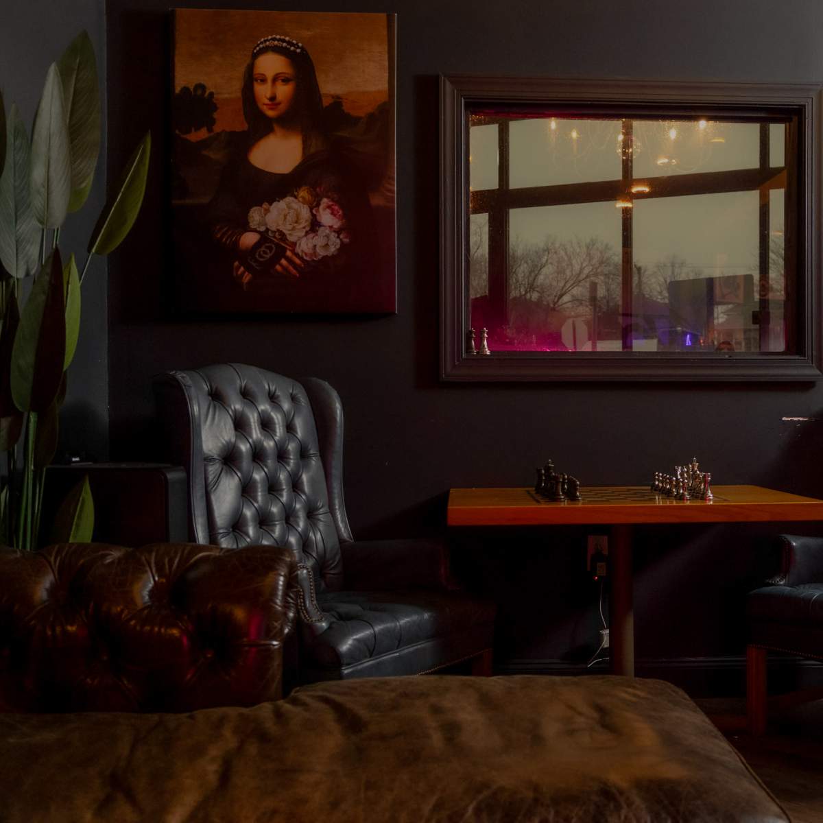 A cozy lounge area with dark walls, featuring a large portrait of a woman, a small wooden table with a chess set, and two tufted leather armchairs.