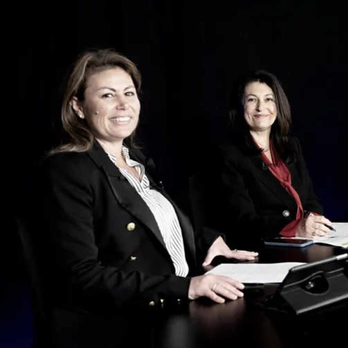 Four individuals sit at a table in a dimly lit studio, facing the camera with documents and devices in front of them.