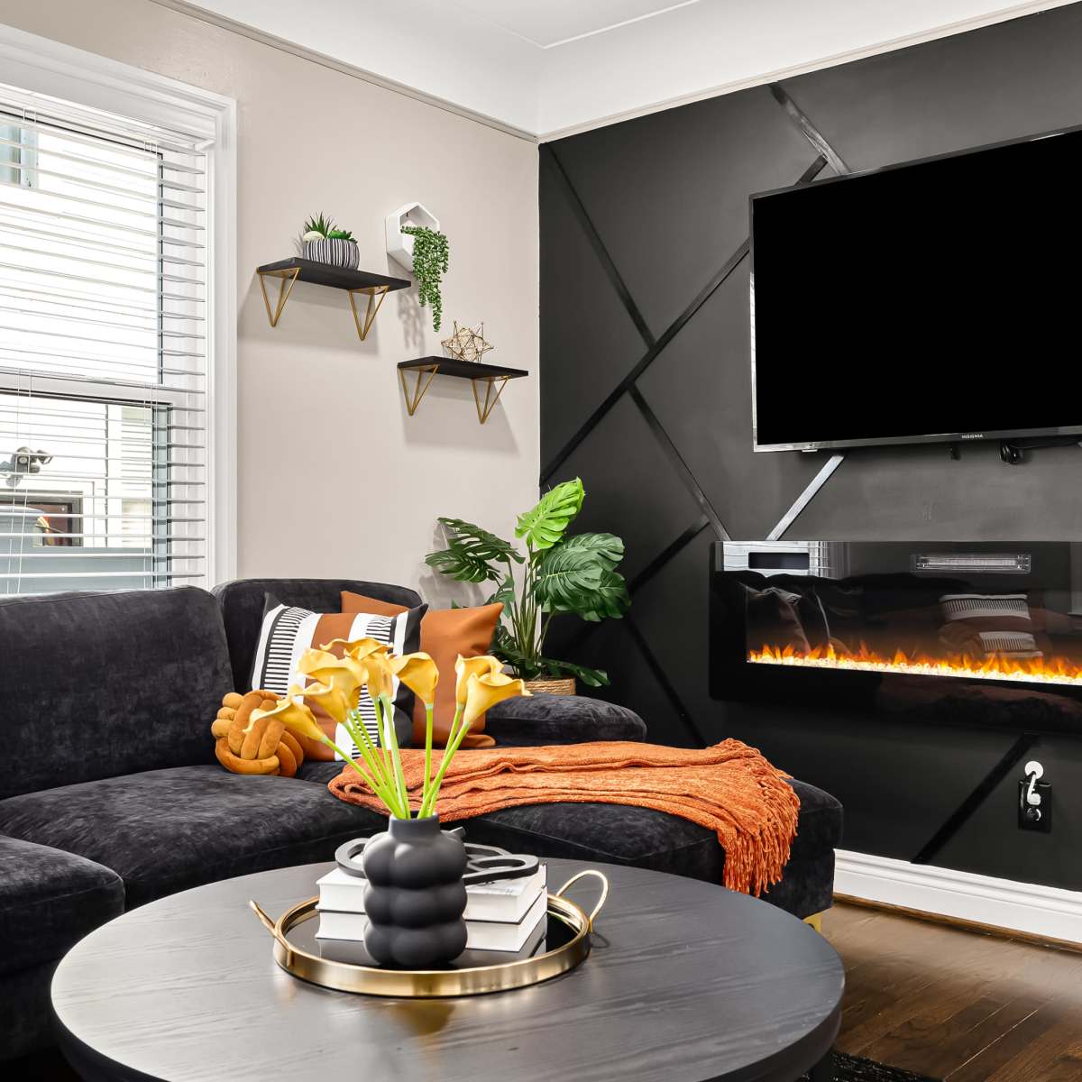 The living room features a dark sectional sofa, a round coffee table, and a mounted TV above an electric fireplace, with decorative plants and shelves on the walls.