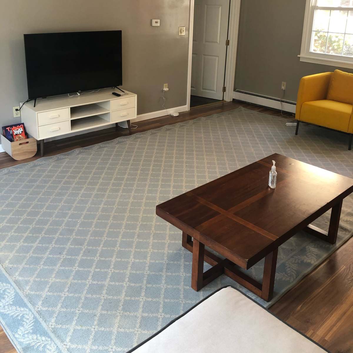 The living room features a television on a white stand, a wooden coffee table, a yellow chair, and a light blue area rug on a hardwood floor.