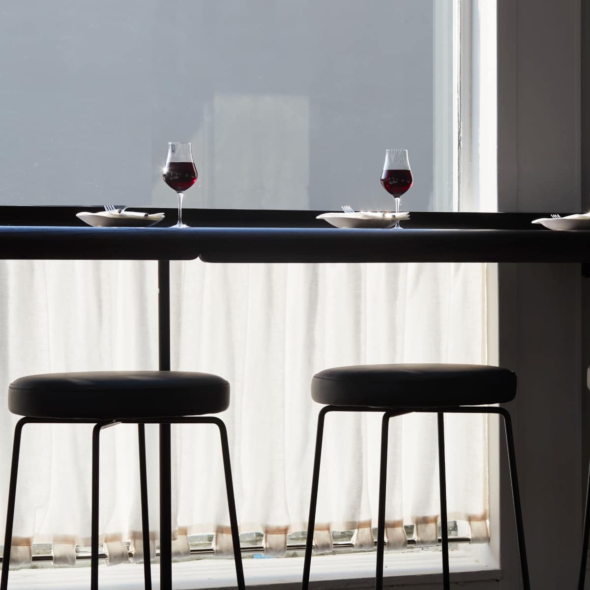 A dimly lit bar area features three black stools with glasses of red wine and small plates on a dark table near a window with sheer curtains.