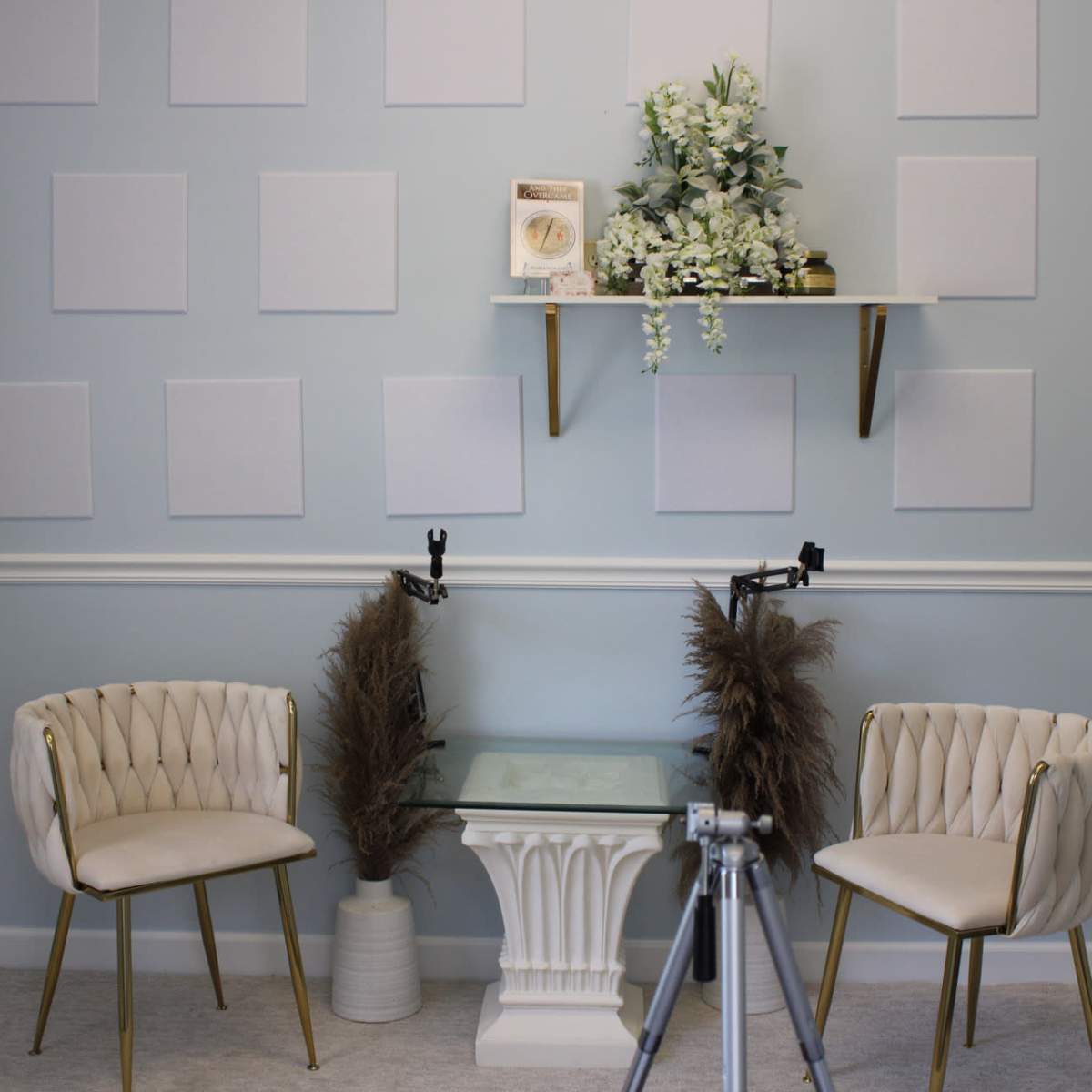 A well-arranged room with light blue walls, two white upholstered chairs with gold legs, a glass table, and decorative elements on a shelf, including plants and a round object.