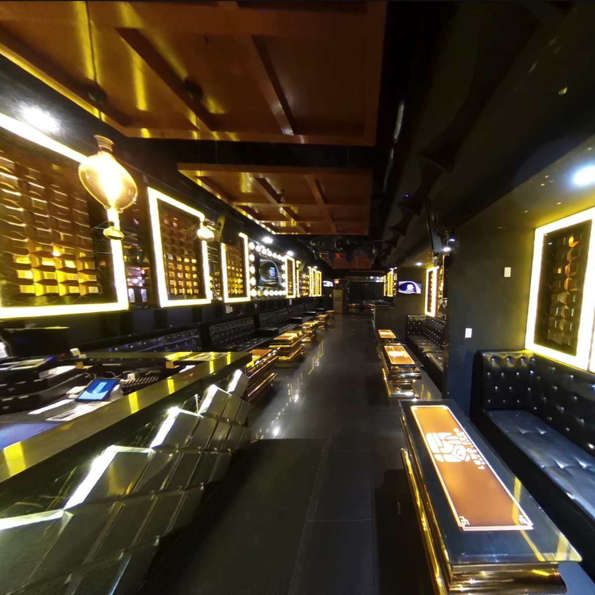 A modern bar interior features a long, sleek counter with illuminated accents and seating along the walls, framed by mirrored panels and warm lighting.