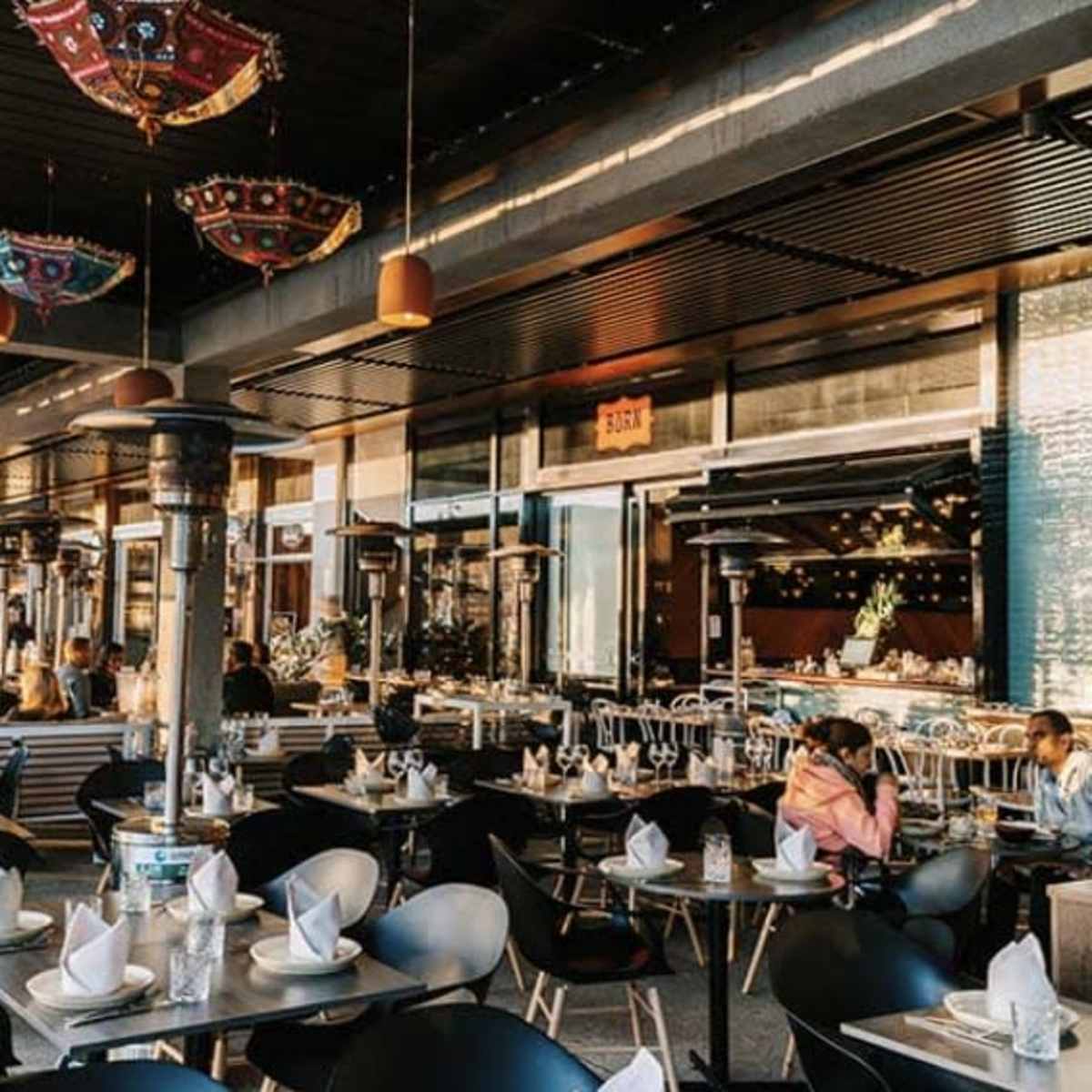 A modern restaurant with outdoor seating, featuring neatly arranged tables and decorative overhead lighting.