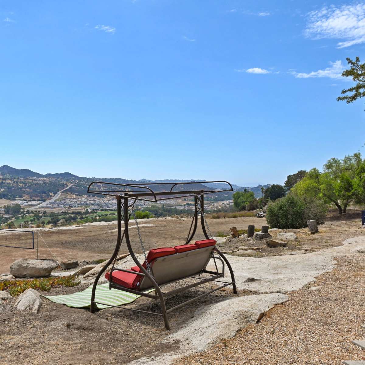 A metal swing chair with red cushions sits on rocky terrain, overlooking a valley and distant mountains under a clear blue sky.