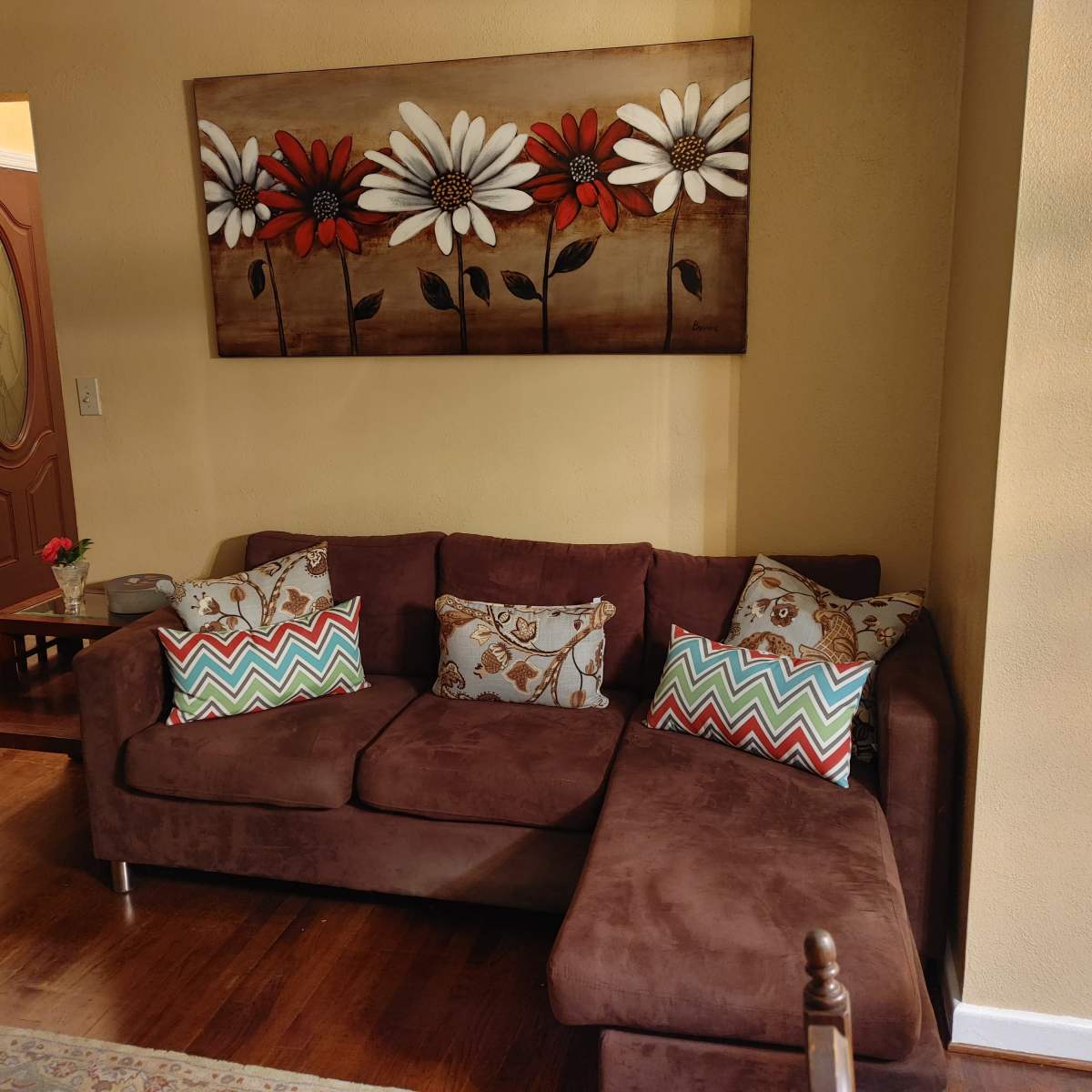 A cozy living room with a brown sectional sofa adorned with colorful patterned pillows, next to a wooden coffee table and a wall decorated with a painting of flowers.