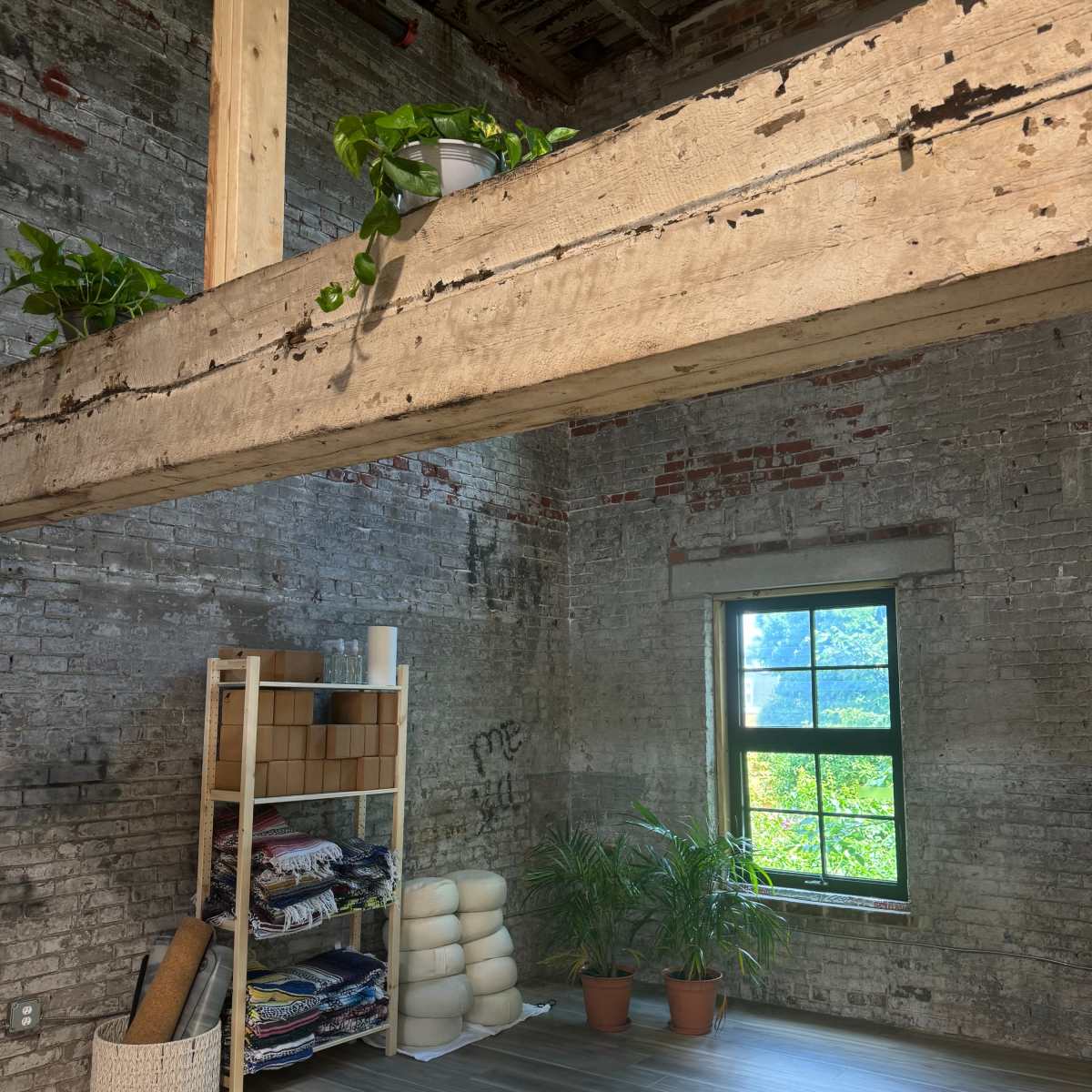 An industrial-style room with exposed brick walls, a wooden beam above, a shelving unit with rolled textiles, and potted plants beside a window.