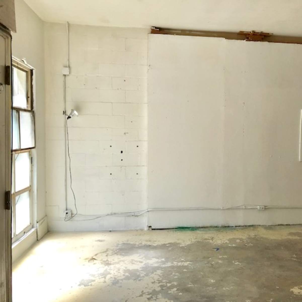 An empty room features a bare white wall with visible marks and a small light fixture attached, along with patchy flooring.
