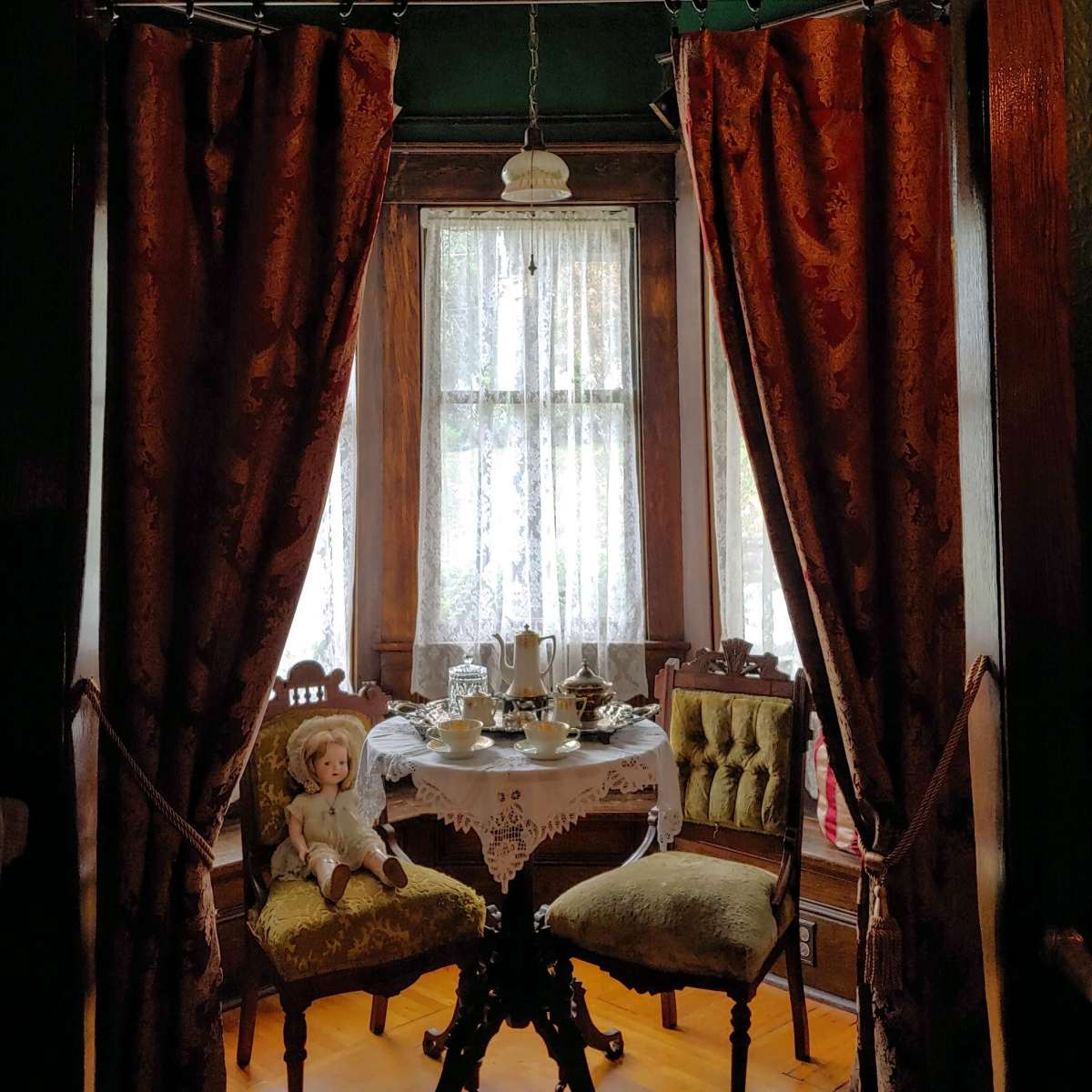 A small vintage dining area is framed by red curtains, featuring a table set for tea and two ornate chairs, with a doll placed on one of the seats.