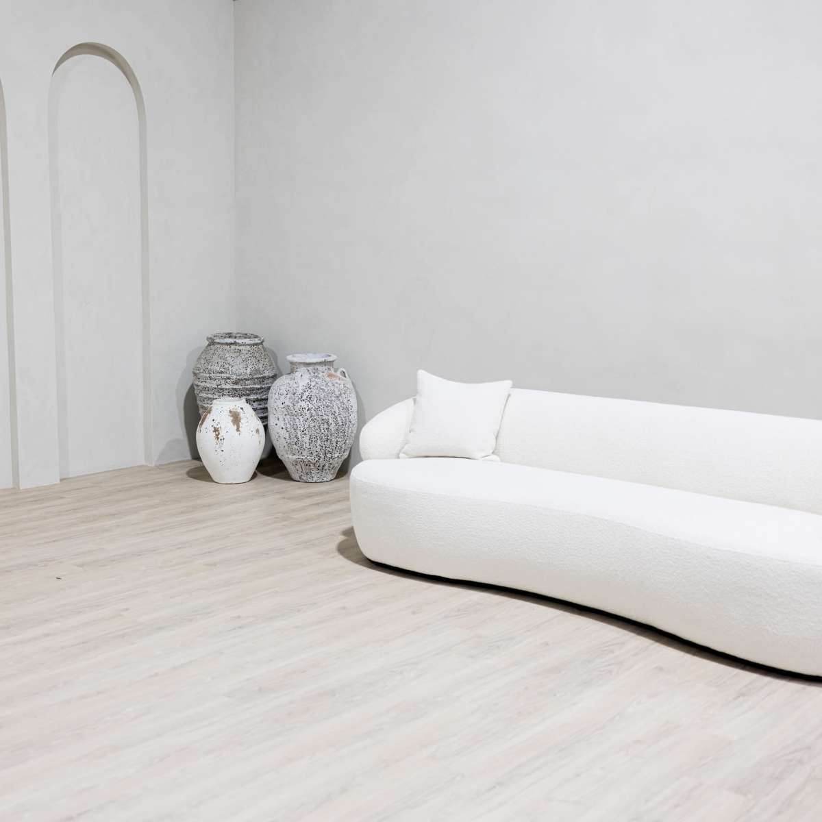 A curved white sofa is positioned next to three decorative vases in a minimalist room with arched wall niches.