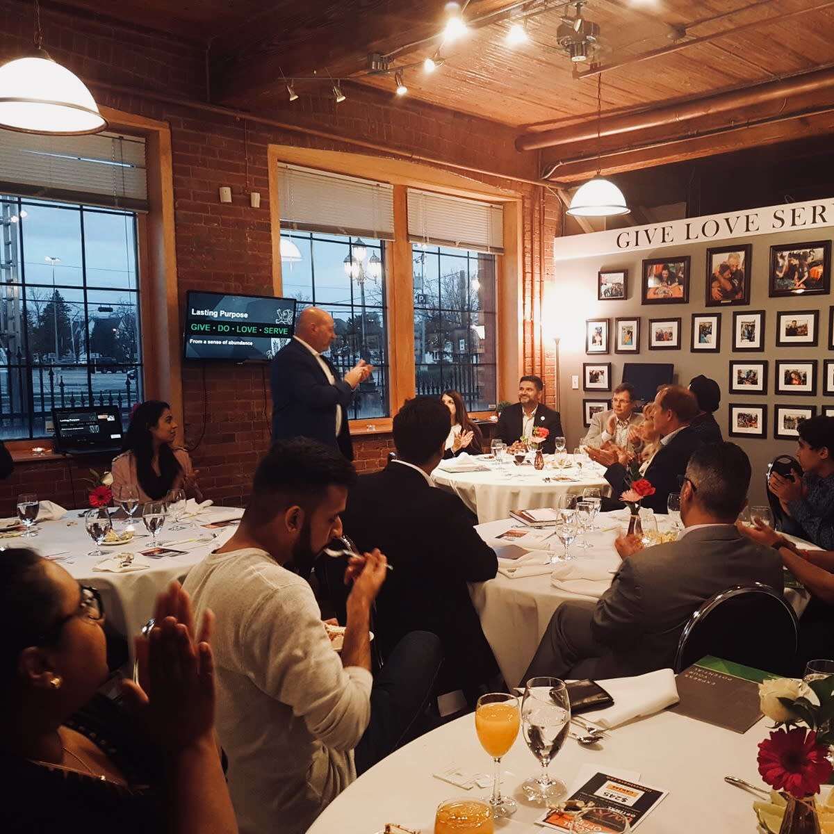 A speaker stands before a seated audience at a dinner event, with tables set and participants engaged in applause.