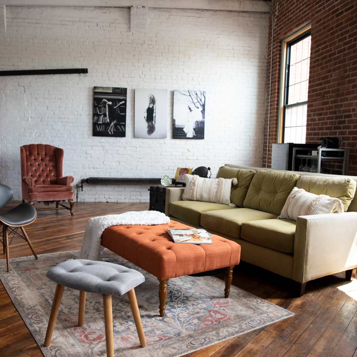A cozy living room featuring a green sofa, an orange upholstered ottoman, a gray stool, and a burgundy armchair, with exposed brick walls and wooden floors.