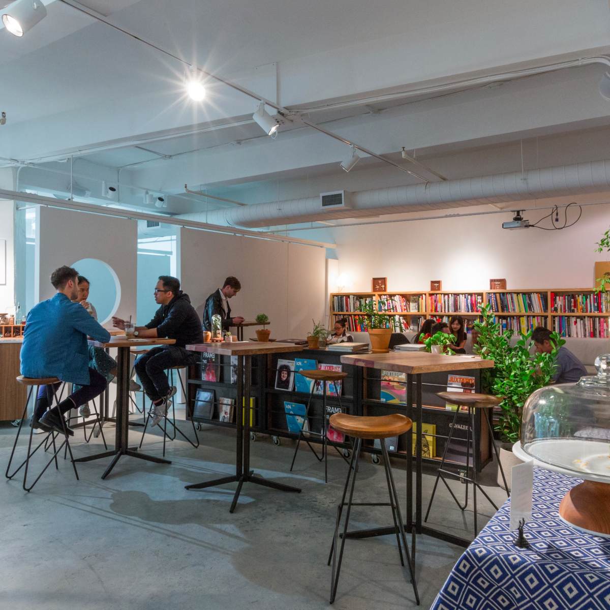 A modern, brightly lit café with patrons seated at high tables, surrounded by bookshelves filled with books and plants.