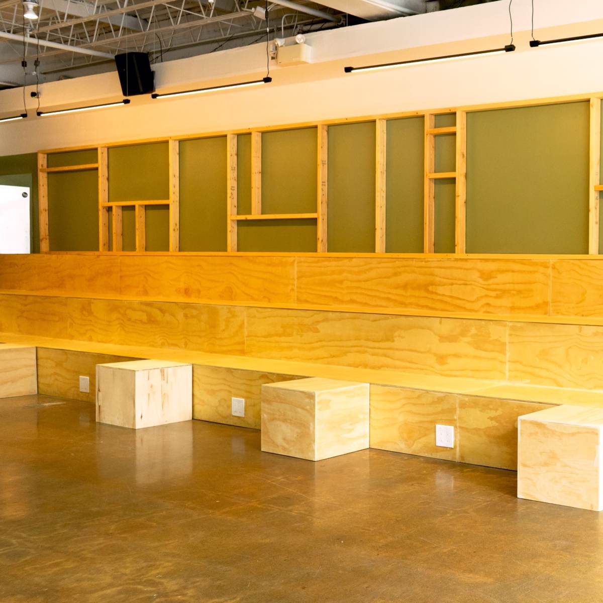 A minimalist interior space with a wooden bench, cuboid seating, and a green accent wall, complemented by a potted plant.