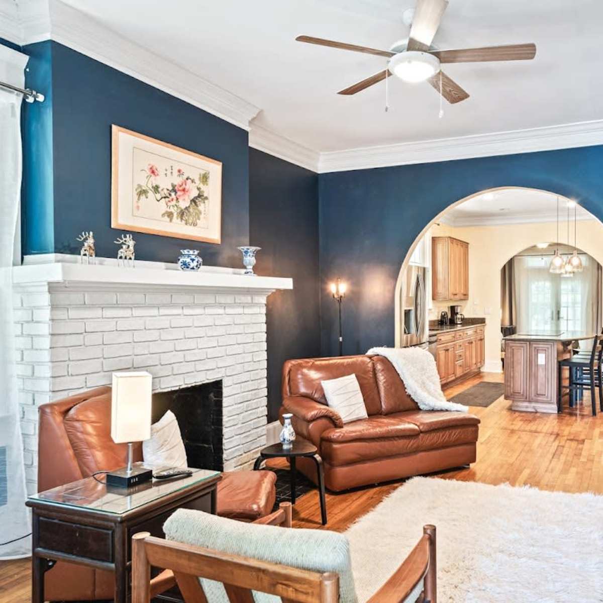 A living room with a brown leather sofa and armchair, a white rug, a decorative fireplace, and an archway leading to a kitchen area.