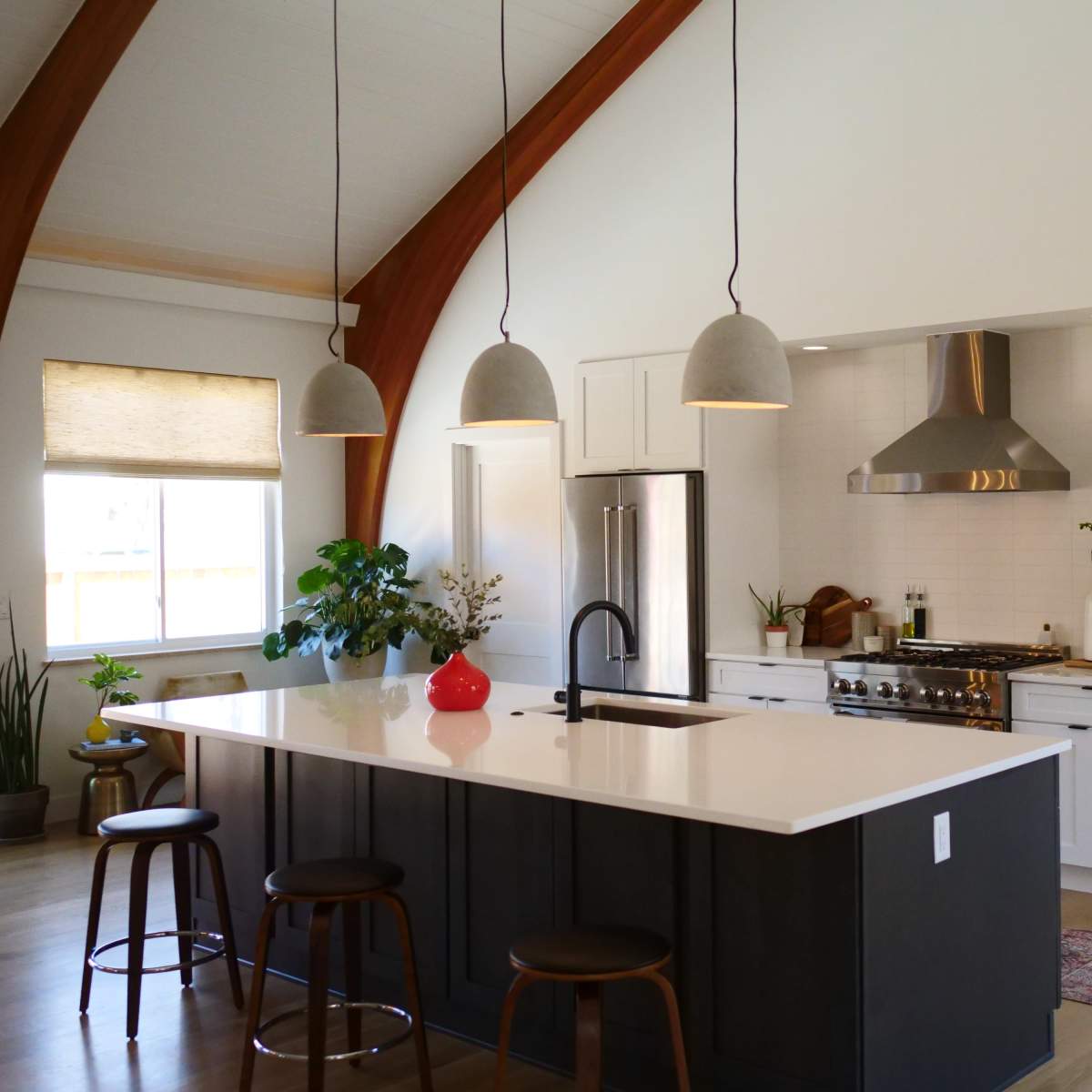 A modern kitchen featuring a large island with bar stools, stainless steel appliances, and pendant lights hanging from a high ceiling with wooden beams.