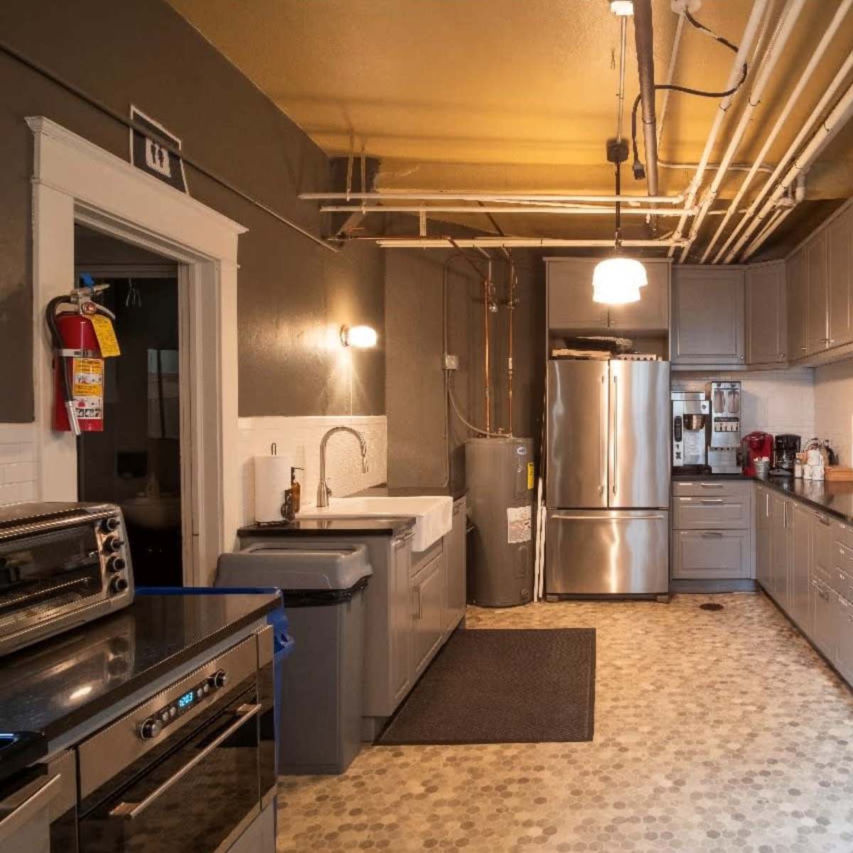 A modern kitchen features gray cabinetry, stainless steel appliances, a large sink, and a black countertop, with visible plumbing pipes overhead.