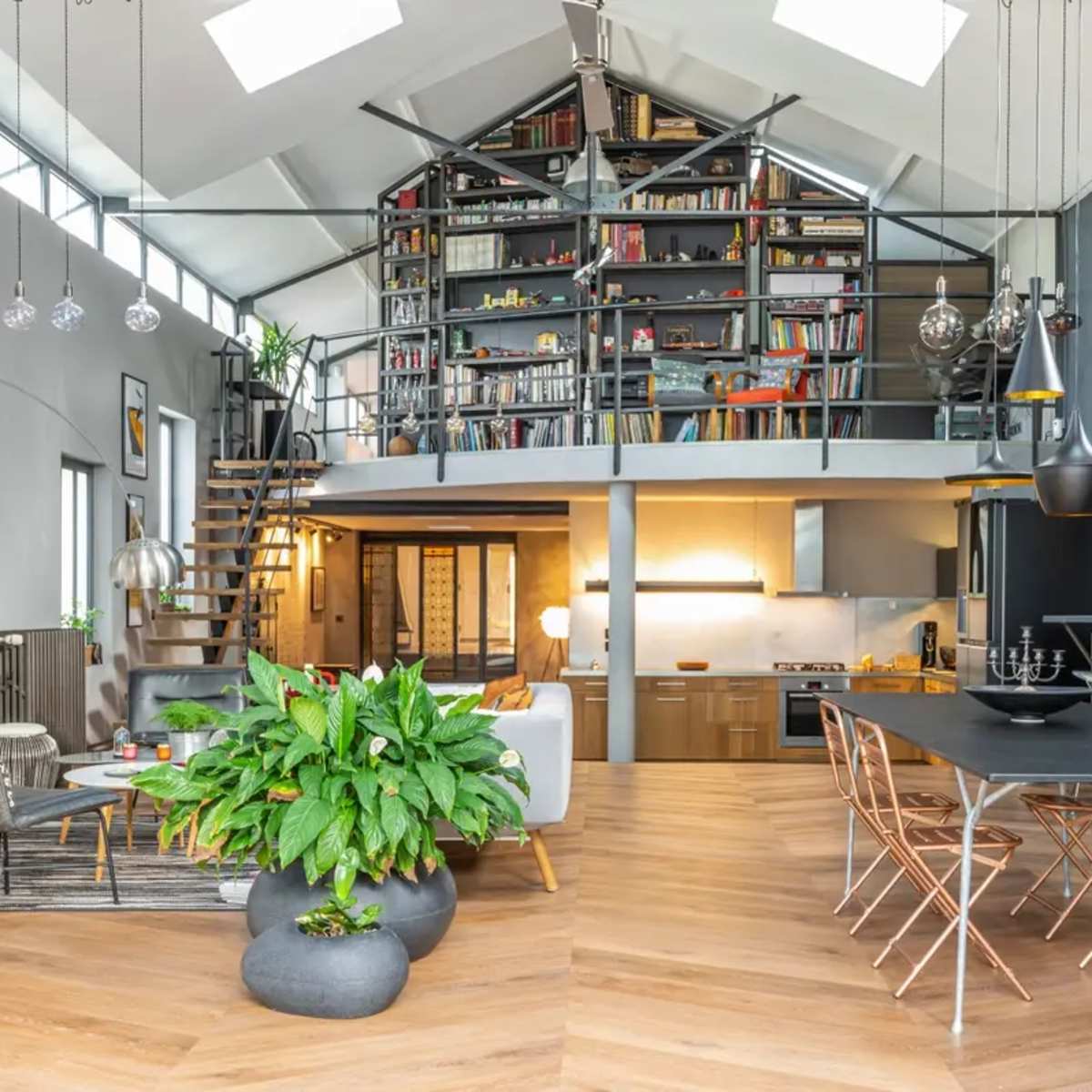 A modern open-concept living space with a dining area, kitchen, and a lofted bookshelf, featuring large windows and wooden flooring.