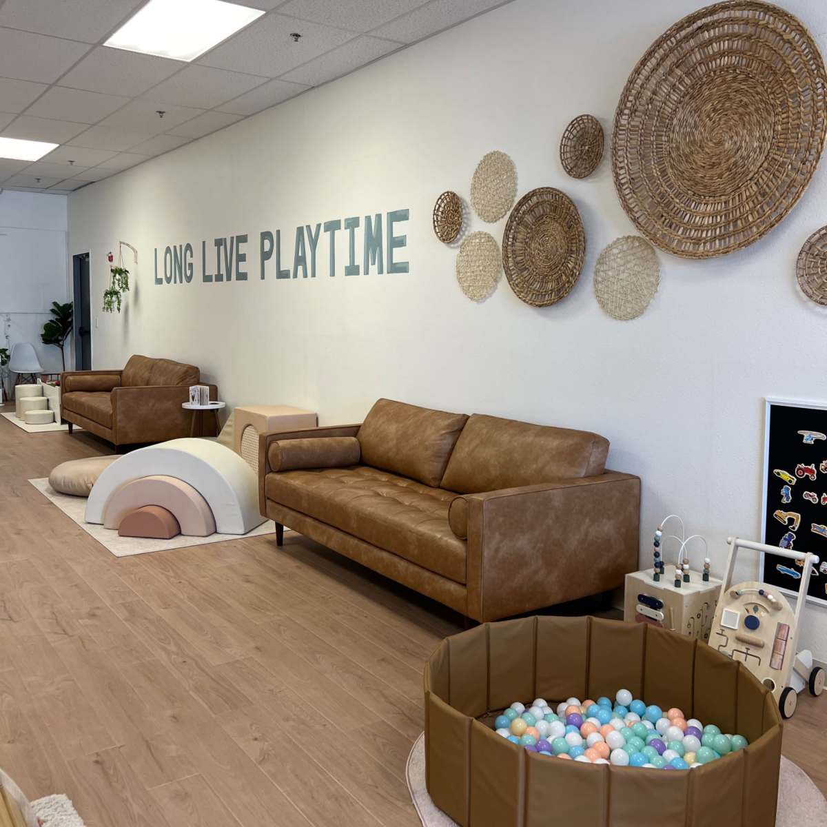 A brightly lit playroom with two brown leather sofas, decorative woven wall hangings, and a large ball pit filled with colorful balls.