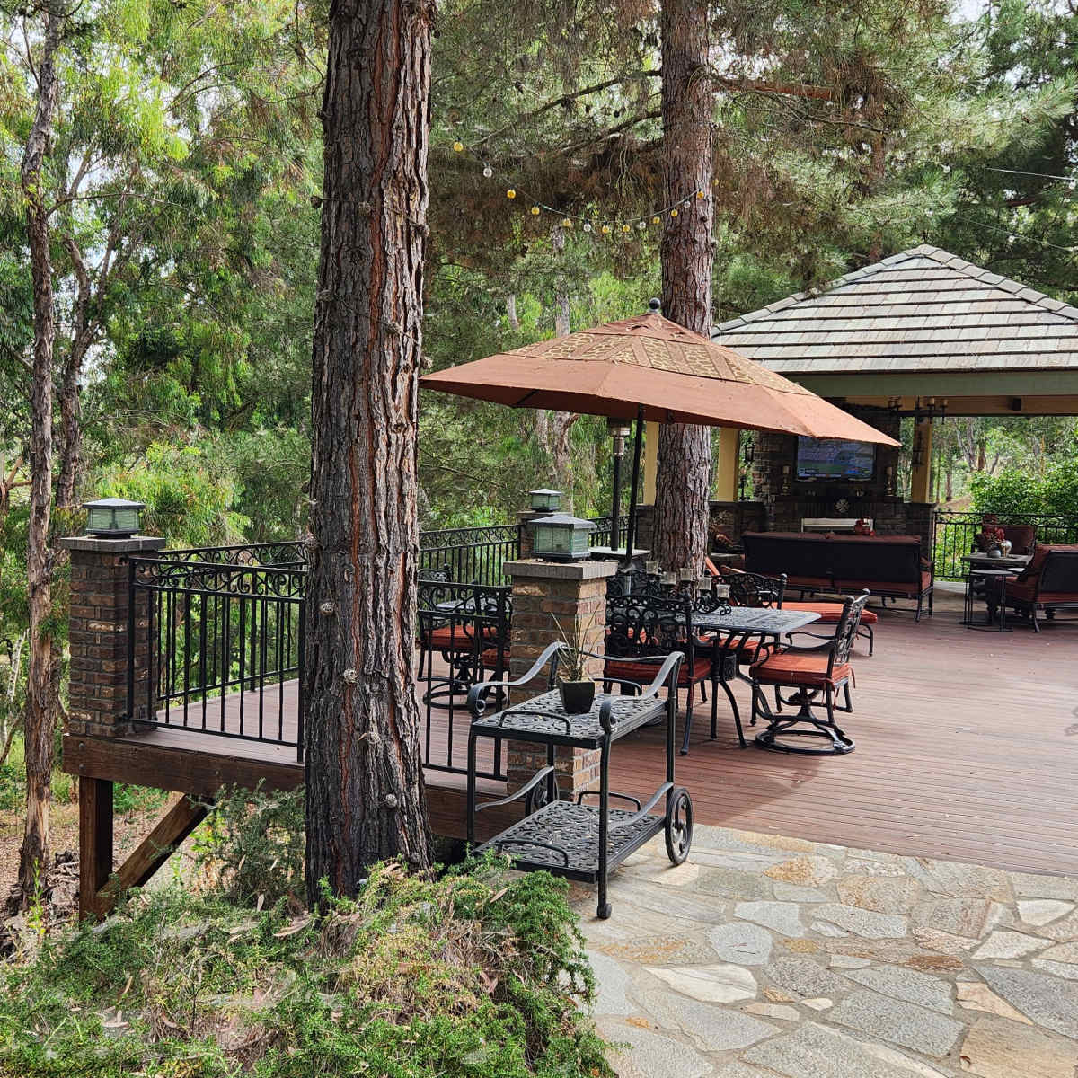 A wooden deck with metal furniture and umbrellas overlooks a forested area, adjacent to a covered seating area.