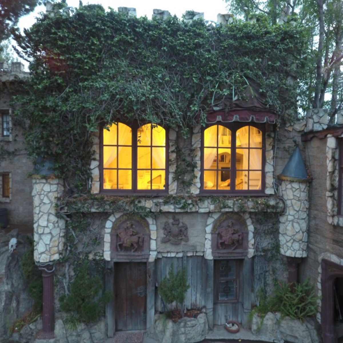 A stone castle-like structure covered in ivy, featuring large windows that emit a warm glow.