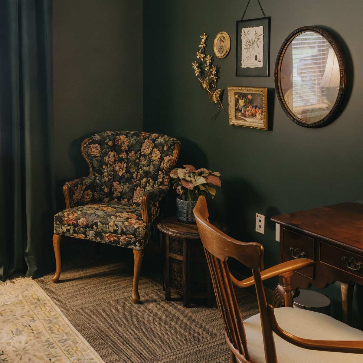 A dark green room with a floral upholstered chair, a wooden desk with a mirror, and a small plant beside a window with curtains.
