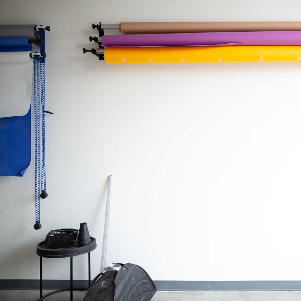 A wall with several rolls of colored backdrop paper mounted on a rack, alongside a black stool and equipment on the floor.
