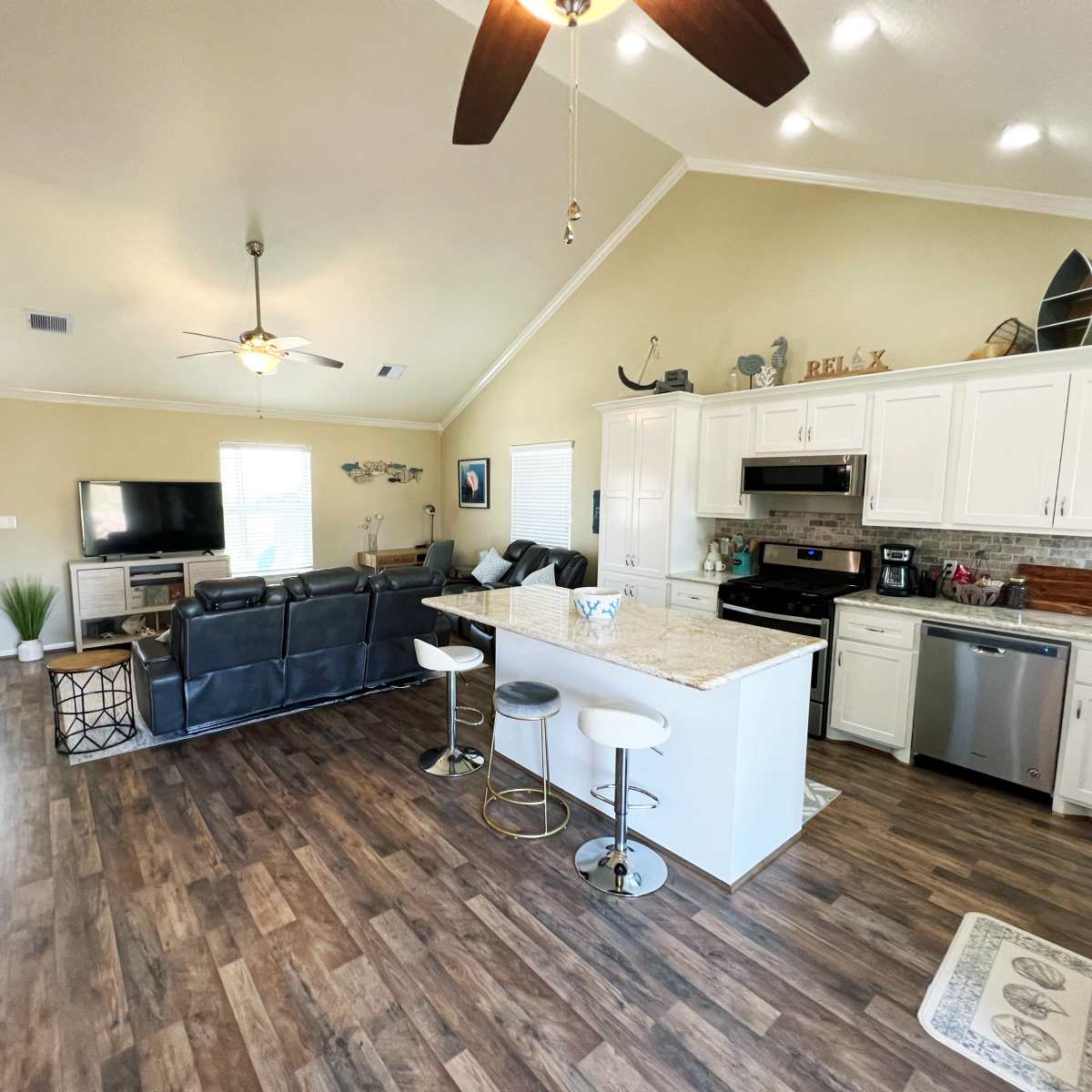 A modern open-concept living area with a kitchen, featuring white cabinets, a central island with seating, and a living room with a large television and seating arrangement.