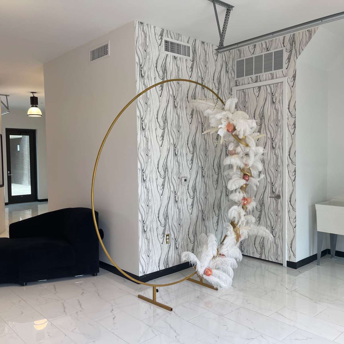 A modern interior space featuring a curved gold sculpture adorned with white fabric and flowers, alongside a dark blue sofa and patterned wallpaper.