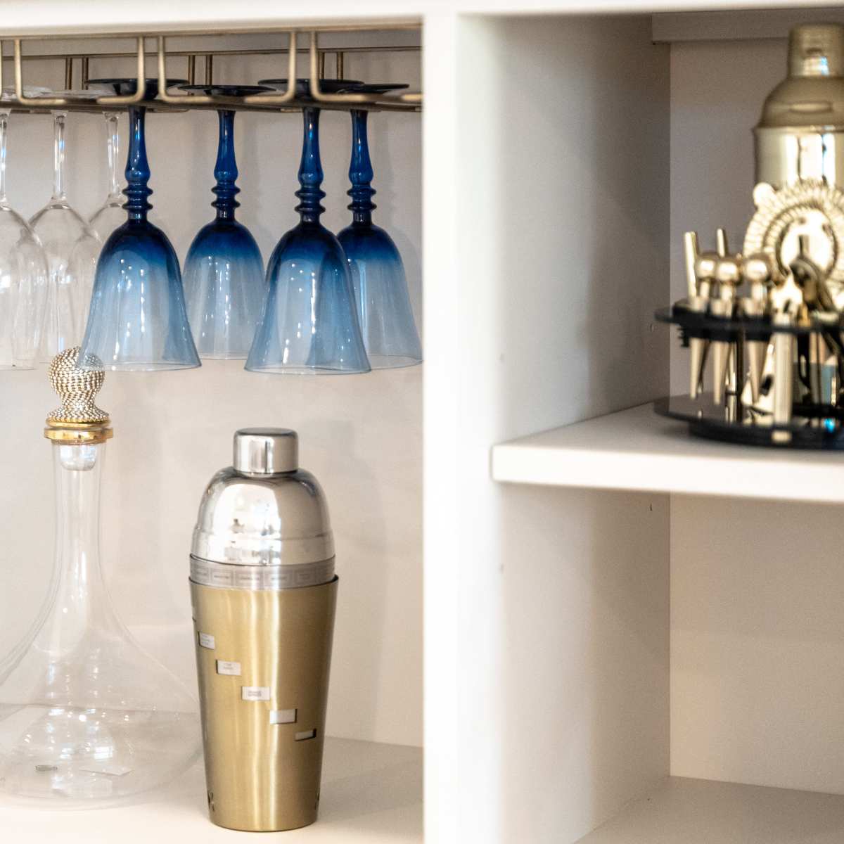 A cabinet interior with hanging wine glasses, a blue-toned collection of stemware, a decanter, a cocktail shaker, and a bar tool set.