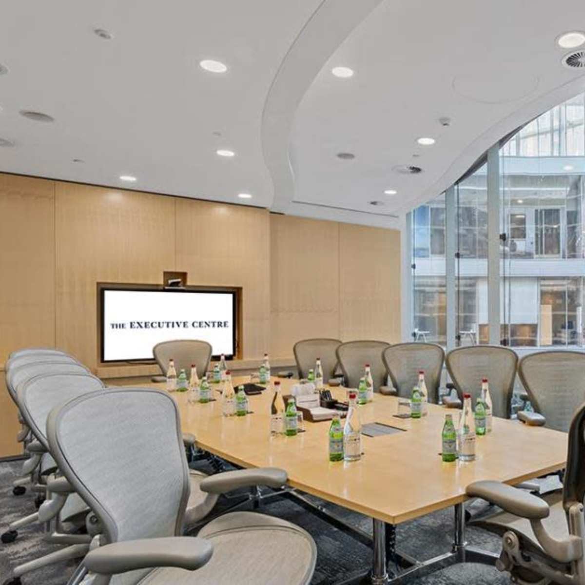 A modern conference room with a large oval table surrounded by ergonomic chairs, a presentation screen displaying "THE EXECUTIVE CENTRE," and large windows offering a view of an atrium area.