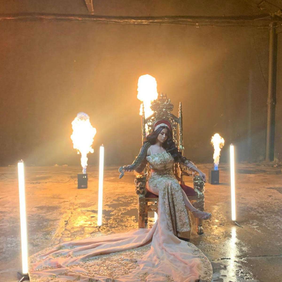 A figure in an ornate gown sits on a throne surrounded by flames and illuminated by spotlights in a dimly lit industrial space.