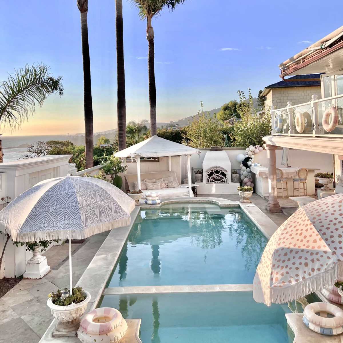 A backyard pool area with palm trees, lounge chairs, and umbrellas, overlooking a scenic view at sunset.