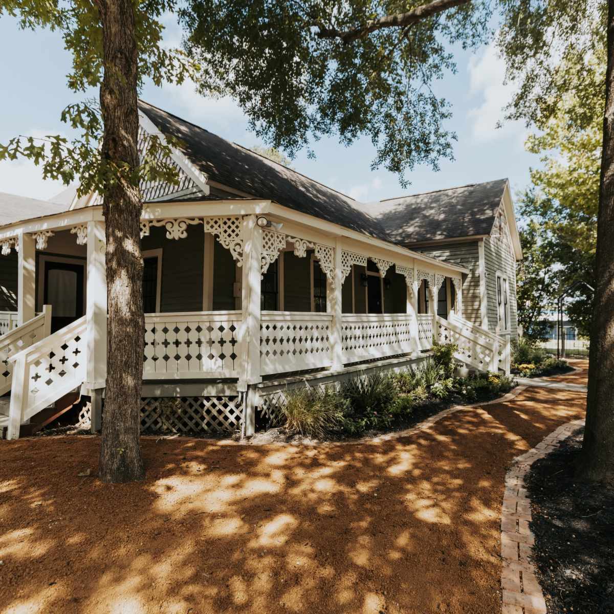 A gray house with a white lattice porch and a gravel path surrounded by trees and landscaping.